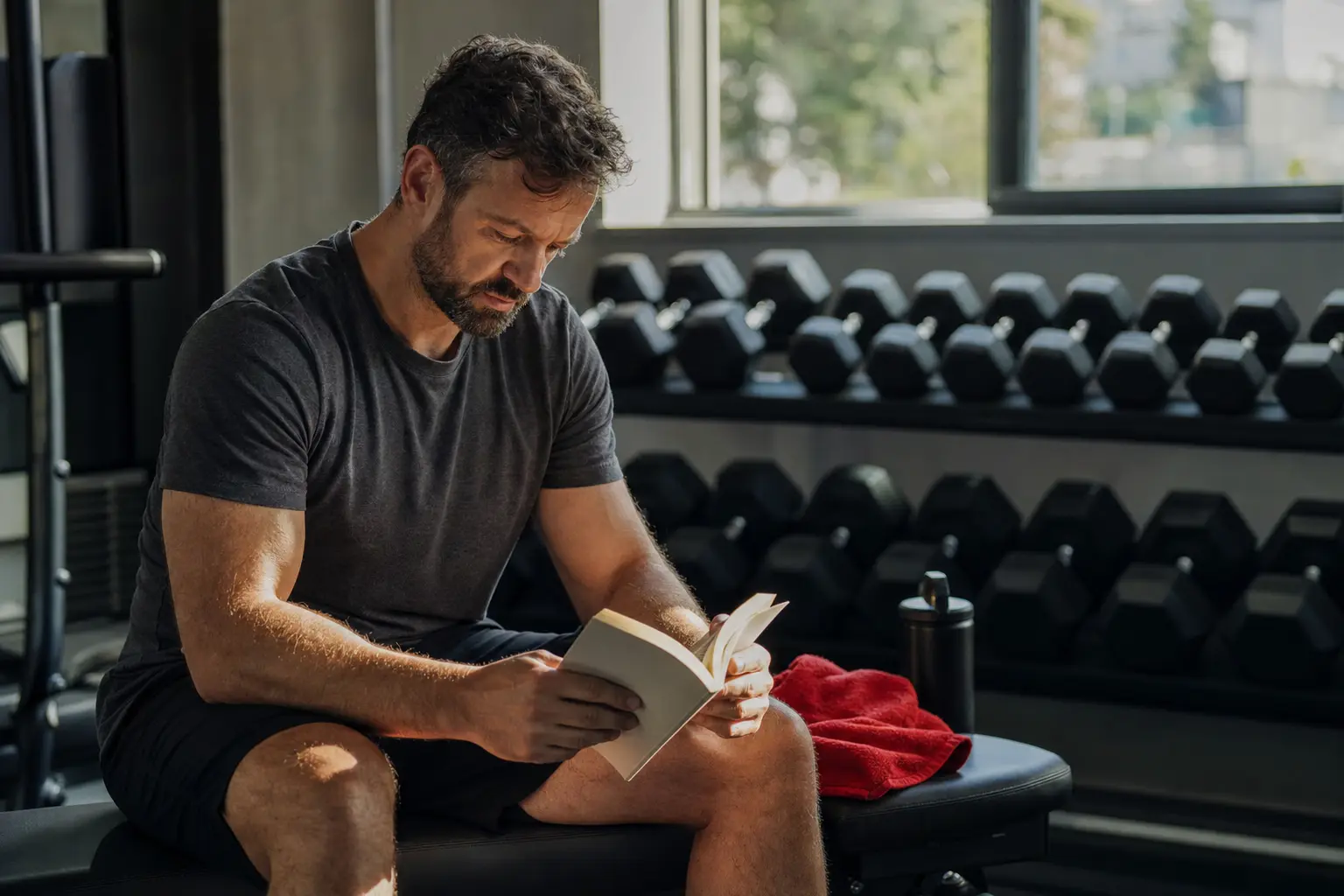 En person som läser på en bänk i ett solbelyst gym mellan seten, med en vattenflaska bredvid sig.