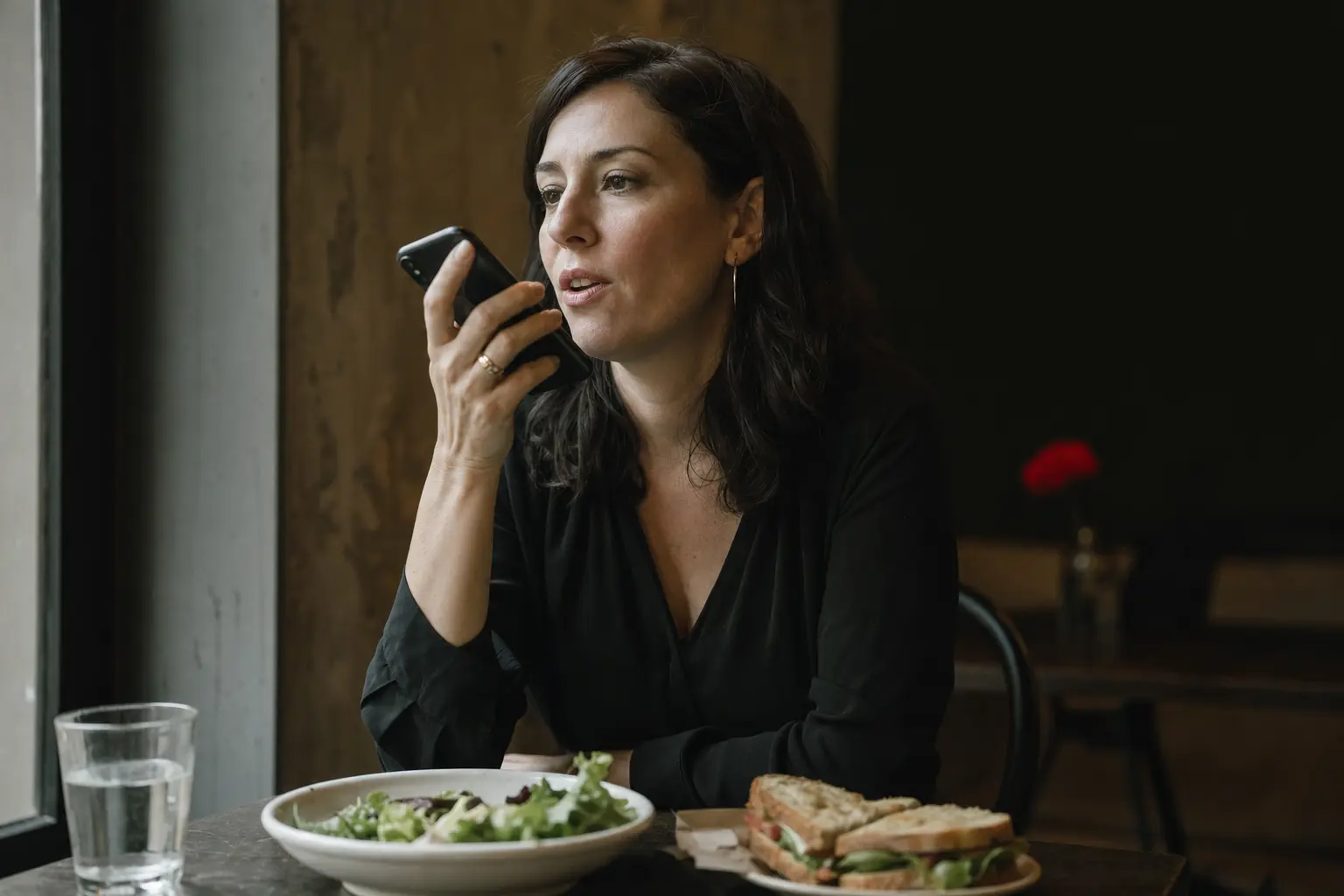 A woman speaking to her phone at a cafe table over lunch.